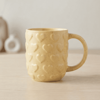 كوب سيراميك أصفر بنقوش قلوب بارزة.  بالإنجليزية: Yellow Ceramic Mug with Embossed Heart Pattern.